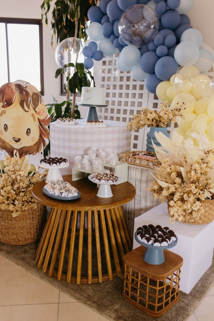 Cozy baby shower table decorated with blue balloons and treats, featuring a cute lion illustration.